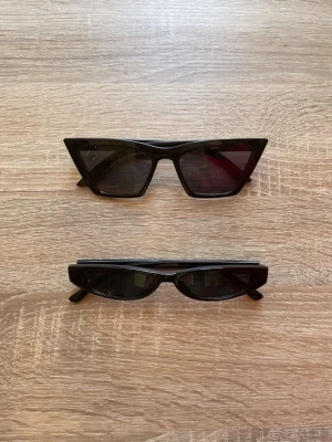 Två svarta solglasögon - Säljer två svarta solglasögon i trendiga modeller. Ena har en markerad cat-eye form och den andra har en smal design. Båda är i svart plast och passar perfekt för en cool och modern stil. Vet ej märket. Använt några gånger. Köp båda för 100kr eller en för 50kr.