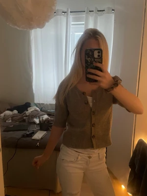 Kortärmad beige kofta med knappar - Säljer en beige kofta med korta ärmar och knappar framtill. Koftan har en croppad passform och ribbstickade detaljer vid ärmslut och nederkant. Perfekt att styla över ett linne eller t-shirt för en chill look.