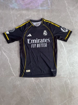 Real Madrid svart matchtröja Adidas - Säljer en svart Real Madrid matchtröja från Adidas med gula detaljer och vita loggor. Tröjan har klubbmärke och sponsortryck på bröstet samt korta ärmar. Tillverkad i ett lätt och ventilerande funktionsmaterial, perfekt för fotbollsträning.