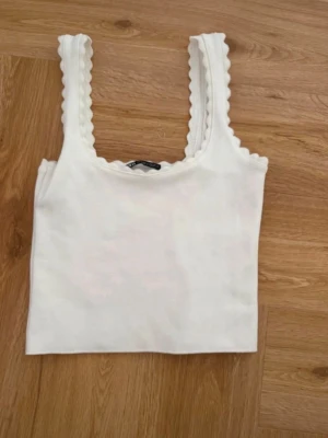 Vit ribbad linnetopp med volangkant - Säljer en söt vit linnetopp med ribbad struktur och volangkant längs axelbanden och ringningen. Toppen är ärmlös och har en croppad passform som passar perfekt till höga jeans eller kjol.