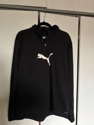 Svart långärmad träningströja Puma - Svart långärmad träningströja från Puma med halv dragkedja och stor vit logga på bröstet. Tillverkad i ett lätt och funktionellt material som passar perfekt för träning.