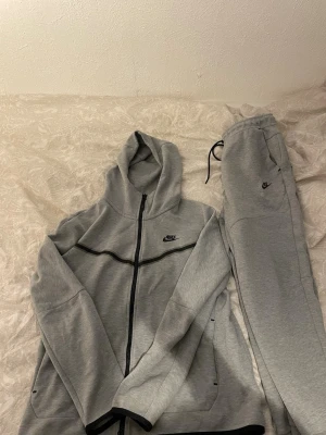 Gråa mjukisbyxor från Nike - Säljer ett par gråa mjukisbyxor och en hoodie  från Nike med dragsko i midjan och fickor på sidorna. Byxorna har en avslappnad passform och är gjorda i mjukt bomullsmaterial. Perfekta för chill dagar eller när du vill vara bekväm.