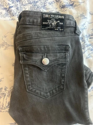 Grå jeans från True Religion - Snygga grå jeans från True Religion med klassisk ficka och silverfärgade knappar. Jeansen har en rak passform och coola sömdetaljer på bakfickan. I bra skick, köpta för 1300kr