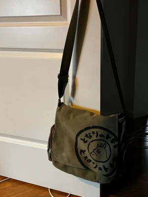 Olivgrön axelväska med Totoro-tryck - Olivgrön axelväska i canvas med justerbar axelrem och stort svart Totoro-tryck på framsidan.  Den är lite trasig via det mindre facket fram men går nog lätt att limma eller sy igen. Allt annat är som de ska vara. 