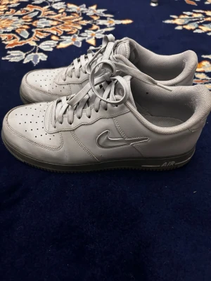Grå Nike Air Force 1 sneakers - Storlek 45. Säljer ett par klassiska grå Nike Air Force 1 sneakers i lågt utförande. Skorna har en stilren design med perforerad tå, snörning och den ikoniska Swoosh-loggan på sidan. Tillverkade i läder med gummisula och AIR-dämpning för extra komfort.