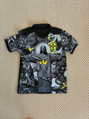 Nike Brasil grafisk fotbollströja - Unik svart och grå fotbollströja från Nike med Brasilien-tema. Tröjan har ett stort grafiskt tryck av Kristusstatyn, CBF-logga och gula detaljer. Korta ärmar och rund hals. Perfekt för dig som vill sticka ut på planen eller heja på Brasilien.
