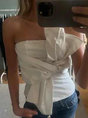Vit axelbandslös topp med stor rosett - Säljer en superfin vit axelbandslös topp med en stor rosett framtill. Toppen har en bandeauformad passning och är perfekt för dig som vill sticka ut. Snygg till både jeans och kjol!