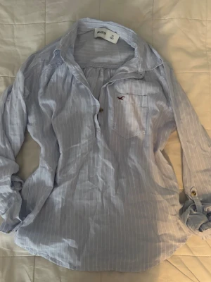 Ljusblå randig skjorta från Hollister - Säljer en ljusblå skjorta från Hollister med tunna vita ränder och en liten röd logga på bröstfickan. Skjortan har lång ärm, klassisk krage och knappar framtill. Perfekt för en avslappnad men ändå stilren look.