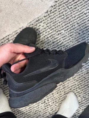 Svarta Nike mesh sneakers - Säljer ett par helsvarta Nike sneakers med mesh-ovandel och diskret swoosh på sidan. Skorna har en chunky sula och klassisk snörning. Perfekta för dig som gillar sportig och clean stil.