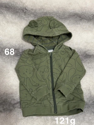 Grön hoodie med djurmönster - Säljer en grön hoodie med dragkedja och huva. Hoodien har ett lekfullt djurmönster med kaniner och katter i en mörkare grön nyans. Perfekt för dig som gillar gulliga detaljer och avslappnad stil.🌸 Fint använt skick. Storlek 68