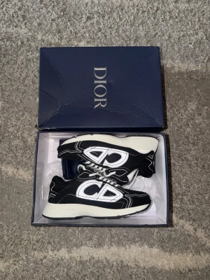 Svarta B30 sneakers - Säljer ett par svarta B30 sneakers med vita detaljer och stora reflekterande loggor på sidorna. Skorna har vit sula, svarta meshpaneler och vita skosnören. Kommer i originalkartong och dustbag. Perfekta för dig som gillar exklusiva och trendiga sneakers.