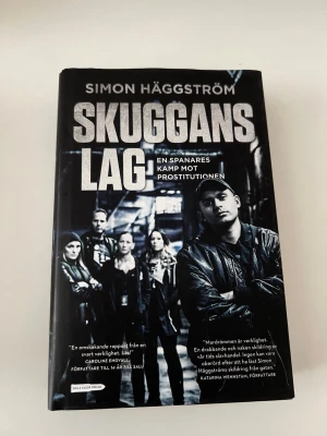 Skuggans lag av Simon Häggström - En svart hårdinbunden bok med titeln 'Skuggans lag' av Simon Häggström. Boken handlar om en spanare och hans kamp mot prostitutionen i Stockholm. Omslaget har ett svartvitt foto av flera personer i en urban miljö. Utgiven av Kalla Kulor Förlag. Mycket bra skick förutom att omslaget har råkats vikas en gång. Med på bild 3. Boken är inbunden. 