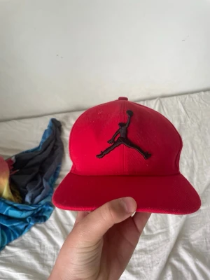 Röd Jordan keps med broderad logga - Snygg röd keps från Jordan med den klassiska Jumpman-loggan broderad i svart framtill. Kepsen har böjd skärm och justerbar baksida. Insidan är fodrad med svarta band där det står 'JORDAN' i vitt. Perfekt för dig som gillar streetwear och sportig stil.