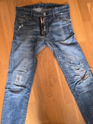 Blå slitna jeans med hål och färgstänk från dsq2 - Säljer ett par orginal blå jeans med skinny fit och tydliga slitningar, hål på knäna och coola färgstänk. Jeansen har klassisk femficksmodell och dragkedja. Perfekta för dig som gillar en avslappnad och trendig streetstil.
