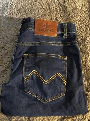 Morris Stevie Satin Jeans blå 29/30 - Mörkblå jeans från Morris i modellen Stevie Satin Jeans. Klassisk femficksdesign med kontrastsömmar i gult och läderpatch bak i midjan. Snygg V-formad söm på bakfickan. Perfekta för dig som gillar stilrena och tidlösa jeans.