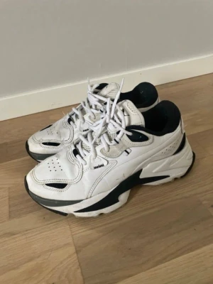 Vita chunky sneakers från Puma - Ett par vita sneakers från Puma med svarta detaljer och en robust sula. Skorna har vita skosnören och Puma-logotypen syns på hälen och sulan. Fint skick, inga hål i hälen men är lite slitet och nedtrampat. Inget som jag känner när jag har på dem dock. Soft foam sula så riktigt sköna
