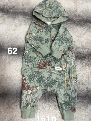 Grön jumpsuit med djungelmönster - Säljer en grön jumpsuit med huva och djungelinspirerat mönster med leoparder, apor och papegojor. Mjuka muddar vid ärmar och ben, dragkedja på sidan och skön passform. Perfekt för dig som gillar coola prints och bekväma plagg.🌸 Använd en gång. Storlek 62