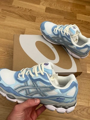 Asics GEL-NYC blåvita sneakers - Snygga sneakers från Asics i vitt och ljusblått med meshpaneler och detaljer i syntet. Skorna har vita snören och en chunky sula i vitt och grått. Perfekta för dig som gillar sportig och trendig stil.