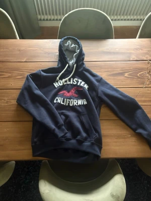 Mörkblå hoodie från Hollister - Mörkblå hoodie från Hollister med vit text och röd logga på bröstet. Stor huva med grått innerfoder och vita dragsnören. Klassisk känguruficka framtill och ribbade muddar vid ärmar och nederkant. Perfekt för en avslappnad och cool stil.