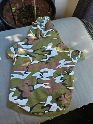 Kamouflagefärgad hundtröja med huva - Hundtröja i grönt, brunt, svart och vitt kamouflagemönster med huva och korta ärmar. Mjukt tyg och ribbade kanter. Perfekt för att ge din hund en cool look på promenaden.