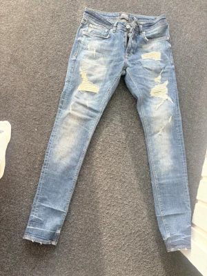 Super dry - Säljer ett par ljusblå skinny jeans med slitningar och hål på både fram- och baksida. Jeansen har klassisk femficksmodell och dragkedjegylf. Perfekta för dig som gillar en avslappnad och trendig stil.