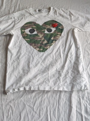 Vit t-shirt med camo-hjärta - Vit t-shirt med stort hjärta i camouflage-mönster och ögon på bröstet. Liten röd hjärtpatch med ögon sitter ovanpå camo-hjärtat. Rund halsringning och korta ärmar. Perfekt för dig som gillar streetwear och unika prints.