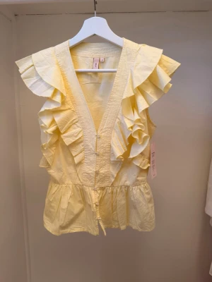 Gul blus med V-ringning - Supersöt gul blus med volanger längs axlar och framstycke. V-ringning och figursydd i midjan ger en feminin siluett. Ärmlös modell i lätt bomullstyg, perfekt för sommardagar. Passar dig som gillar romantisk och trendig stil. Prislapp kvar på, därmed aldrig använd. Hör gärna av er vid frågor💗