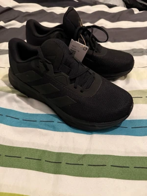 Svarta Adidas sneakers i mesh - Snygga helsvarta sneakers från Adidas med klassiska tre ränder på sidan. Ovandelen är i meshmaterial för bra andningsförmåga och skon har en låg profil med bekväm sula. Perfekta för dig som gillar stilrena och sportiga skor. Helt nya med tag!