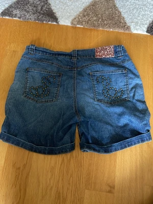 Blå jeansshorts med nitar - Snygga blå jeansshorts med dekorativa nitar i mönster på bakfickorna och en unik detalj med leopardmönstrad patch vid linningen. Klassisk femficksmodell och uppvikta benslut. Perfekta för en avslappnad stil.