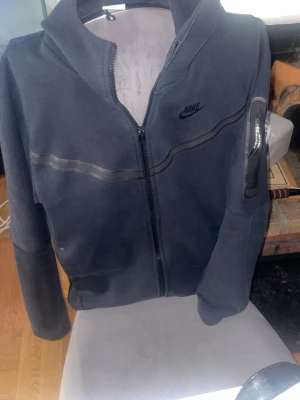 Svart Nike hoodie med dragkedja - Säljer en svart hoodie från Nike med dragkedja framtill och en diskret logga på bröstet. Hoodien har en huva, en ficka med dragkedja på ärmen och en snygg detalj i form av en vågig linje över bröstet. Perfekt för en sportig och avslappnad stil.
