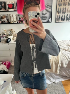 Grå cardigan med knytband - Trendig grå cardigan med långa ärmar och tre knytband framtill. Modellen är rak och har en avslappnad passform, perfekt att slänga över ett linne eller t-shirt. Materialet känns mjukt och bekvämt, och färgen är lätt att matcha med olika outfits.
