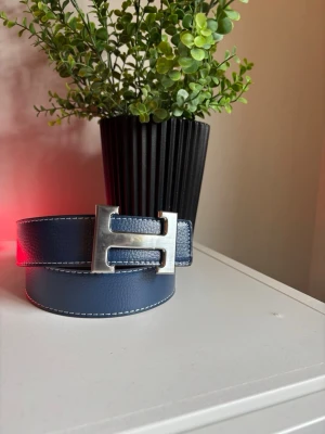 Blått läderbälte med silverfärgat spänne - Snyggt blått bälte i läder med ett stort, silverfärgat H-format spänne. Bältet har vita sömmar längs kanterna som ger en clean kontrast. Perfekt accessoar för att lyfta din outfit och ge en exklusiv touch.