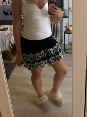 Svart kortkjol med vit spetsvolang - Supersöt svart kortkjol med dubbla lager volang och detaljerad vit spets längs kanterna. Kjolen har en feminin och lekfull look som passar perfekt till sommaren. Elastisk midja för skön passform.