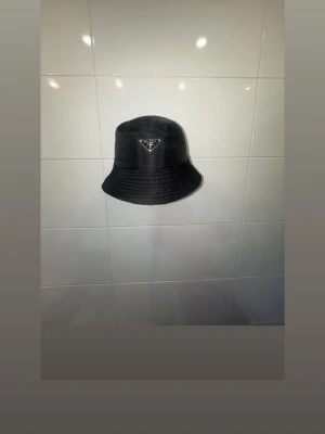 Svart bucket hat - Snygg svart bucket hat med den ikoniska metalltriangeln framtill. Hatten är tillverkad i ett glansigt nylonmaterial som ger en modern och clean look. Perfekt accessoar för att lyfta din outfit och skydda mot solen.