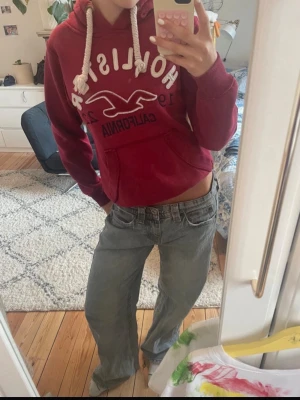 Hollister hoodie - Säljer en vinröd hoodie från Hollister med broderad logga och texten 'California 1922' på bröstet. Hoodien har en stor ficka framtill, vita tjocka dragsnören och mjukt material. Perfekt för chill dagar och snygg till jeans.