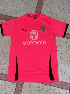 Rosa Marocko fotbollströja Puma - Snygg rosa fotbollströja från Puma med Marockos landslagsemblem på bröstet och trycket 'MOROCCO' framtill. Tröjan har svarta detaljer på ärmar och sidor samt rund hals. Tillverkad i ett lätt och ventilerande funktionsmaterial, perfekt för träning eller match.