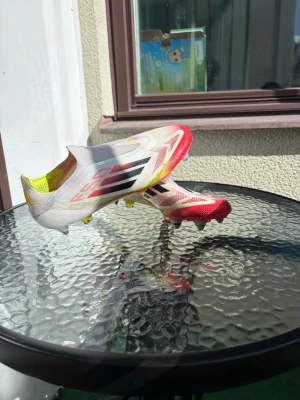 Vita och röda Adidas F50 fotbollsskor laceless - Säljer ett par Adidas F50 fotbollsskor i vitt med röda och gula detaljer samt svarta ränder på sidan. Skorna har en strömlinjeformad design utan snörning och är tillverkade i lätt syntetmaterial för maximal fart på planen.