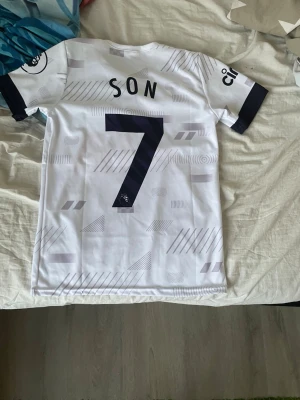 Tottenham Hotspur matchtröja Son 7 - Säljer en vit Tottenham Hotspur matchtröja med Son och nummer 7 på ryggen. Tröjan har svarta detaljer på ärmarna, klubbmärke på bröstet och AIA som sponsortryck. Tillverkad i ett lätt och ventilerande material, perfekt för fotboll eller träning.