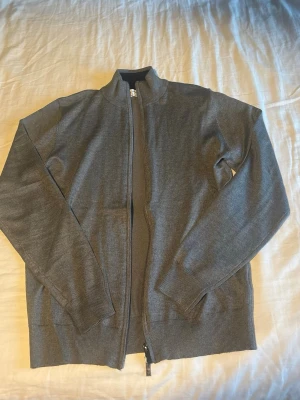 Grå full-zip av premium kashmir och merinoullströja.  - En stilren grå ziptröja från Max Martini från Milano, med ståkrage och långa ärmar. Tröjan har ribbade muddar vid ärmslut och nederkant samt dragkedja framtill. Tillverkad i ett tunt, mjukt premium kashmir och merinoulls material som passar perfekt till lager på lager. (Har storlek L men är något liten i storleken). Nytt skick, aldrig använts.