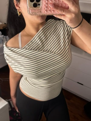 Randig offshoulder topp - Säljer en vit och svart randig offshoulder topp med ribbad struktur. Toppen har en lösare passform upptill och smiter åt i midjan, vilket ger en snygg siluett. Perfekt för dig som gillar en trendig och avslappnad stil.