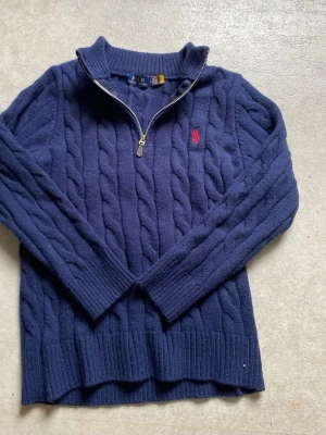 Mörkblå kabelstickad tröja Polo Ralph Lauren - Mörkblå kabelstickad tröja från Polo Ralph Lauren med halv dragkedja och ribbade muddar. Tröjan har en klassisk röd logga broderad på bröstet och hög krage. Perfekt för lager på lager och riktigt snygg till jeans. Hör gärna av er vid funderingar. Priset är inte hugget i sten