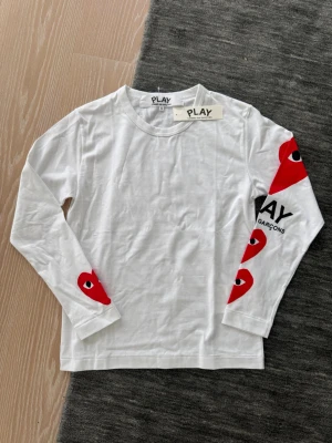 Vit långärmad topp Comme des Garçons - Vit långärmad topp från Comme des Garçons PLAY med röda hjärtmotiv och tryck på ena ärmen. Tillverkad i mjuk bomull och har en klassisk rund halsringning. Perfekt för dig som gillar streetstyle och vill sticka ut med coola detaljer.