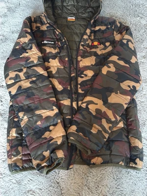Camo pufferjacka från Ellesse - Säljer en camouflagemönstrad pufferjacka från Ellesse med huva och dragkedja framtill. Jackan har vadderad insida och elastiska muddar vid ärmsluten. Perfekt för kalla vinterdagar och riktigt snygg streetstil.