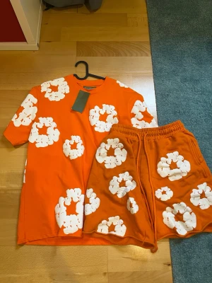 denim tears set orange - Säljer ett färgstarkt set med en orange t-shirt och matchande shorts, båda dekorerade med stora vita blommönster. T-shirten har rund hals och korta ärmar, shortsen har resår i midjan för skön passform. Perfekt för dig som gillar att sticka ut!