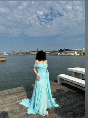 Ljusblå offshoulder långklänning - Säljer en elegant ljusblå långklänning med offshoulder-design och svepande kjol. Klänningen har draperade detaljer över bysten och axelband som knyts i ryggen. Perfekt för speciella tillfällen och ger en romantisk look.