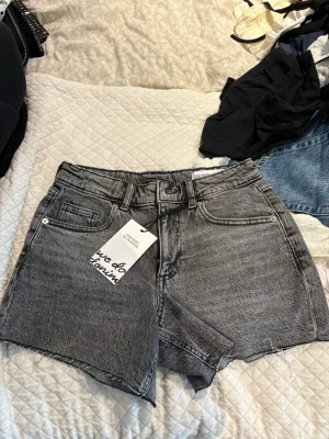 Grå denimshorts med rå kant - Säljer ett par grå denimshorts med hög midja och rå, fransig kant. Shortsen har klassisk femficksdesign, bälteshällor och knappgylf. Perfekta för en avslappnad och trendig look.