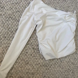 Vit oneshoulder topp med rynk - Säljer en stilren vit oneshoulder topp med lång ärm och snygga rynkdetaljer på sidan. Toppen har en tight passform och är tillverkad i ett mjukt, stretchigt material .