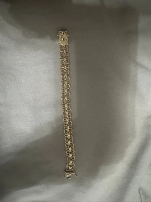 Bred guldlänk armband - Snyggt armband i guldfärgad metall med bred, flätad länkdesign. Armbandet har ett dekorativt lås och en unik struktur som ger ett exklusivt intryck. Perfekt för dig som vill ha ett statement-smycke som sticker ut. Den är guldpläterad.