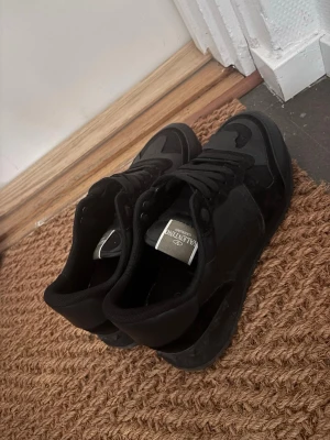 Svarta sneakers från Valentino - Snygga helsvarta sneakers från Valentino med matta och blanka detaljer. Skorna har klassisk låg profil, svarta skosnören och en robust sula. Materialmix av mocka och textil ger en exklusiv känsla och modern look.