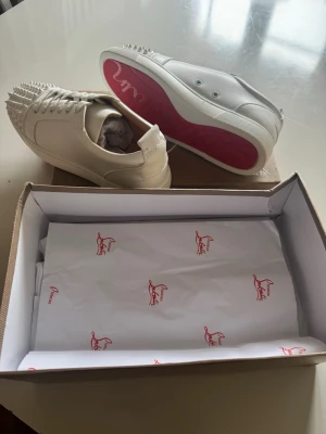 Beige sneakers med nitar och röd sula - Snygga beige sneakers med coola nitar på tån och klassisk röd sula. Skorna har en låg profil och är tillverkade i slätt läder. Perfekta för dig som vill sticka ut med en unik detalj på fötterna.